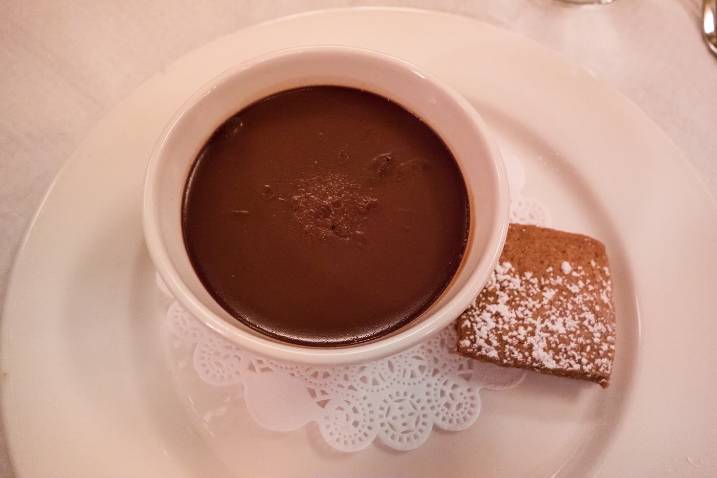 A white ramekin of dark chocolate pot de crème on a doily, with a sugared shortbread alongside